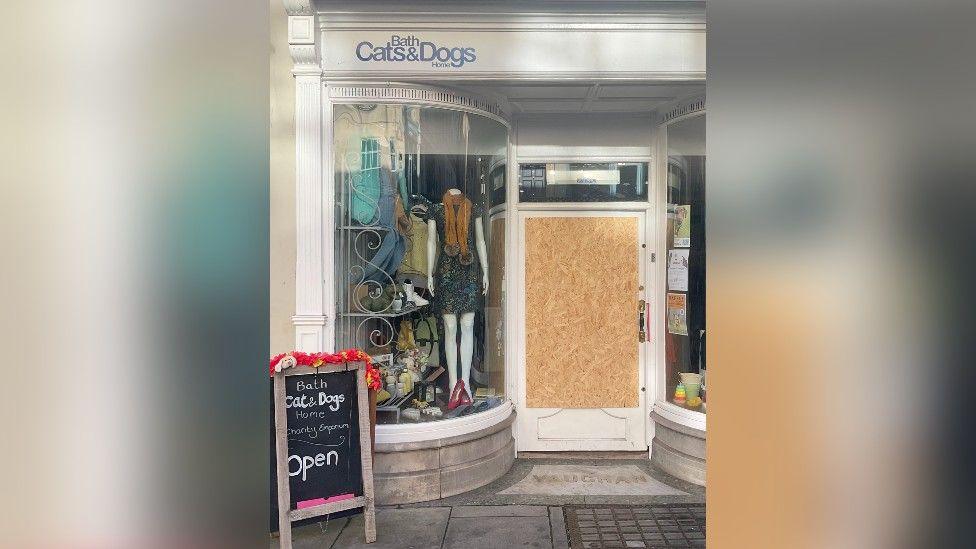 Bath Cats and Dogs Home shop on George Street in Bath. The door of the shop is boarded up with a large piece of wood. Outside there is a A-Frame notice board which says 'Bath Cats and Dogs Home charity shop Open'. 