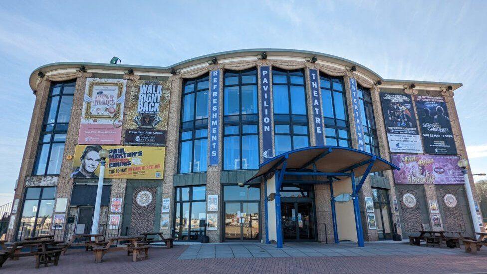 Building with curved entrance and posters of shows on its walls - wooden picnic tables are set outsite its foyer.