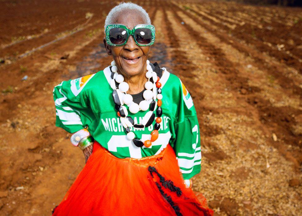 Margret Chola smiling in a field with her hand on her hip, wearing big green bejewelled sunglasses, a green sports jersey, a red dress and big plastic white, red and black beads around her neck.