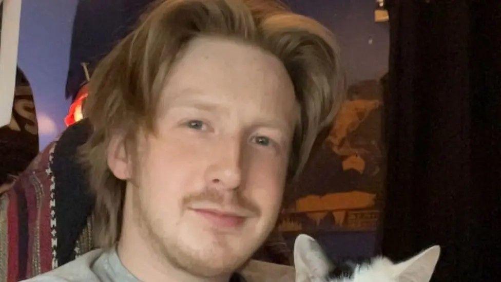 Aaron Smith has long blond hair, parted in the middle and facial hair. He appears to be holding a black and white kitten.