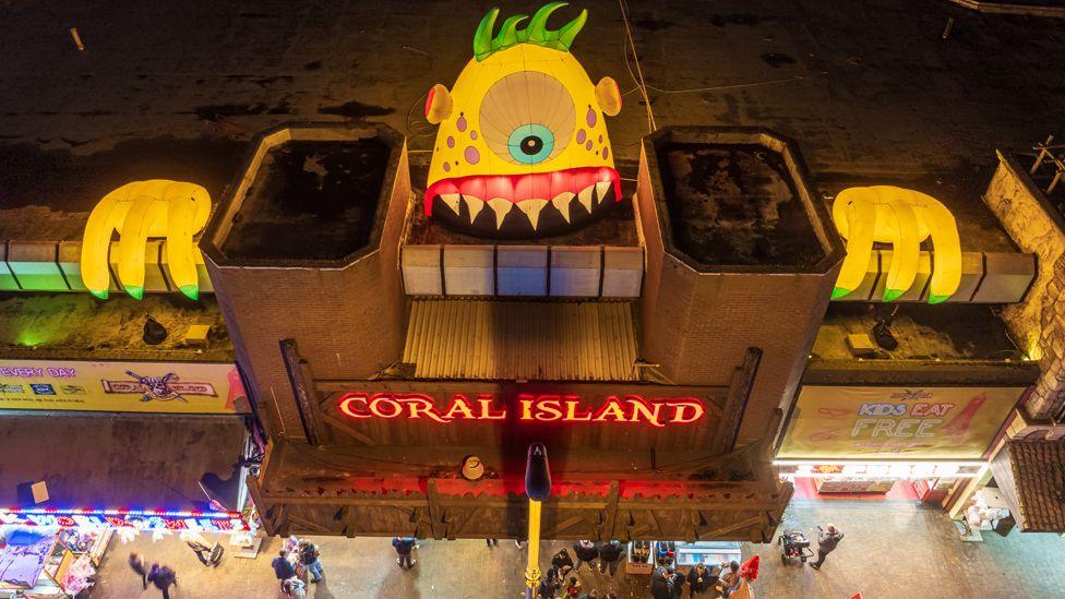 A yellow inflatable monster with spiky teeth, red lips and long yellow fingers peers over the side of the Coral Island amusement arcade, as people walk by below