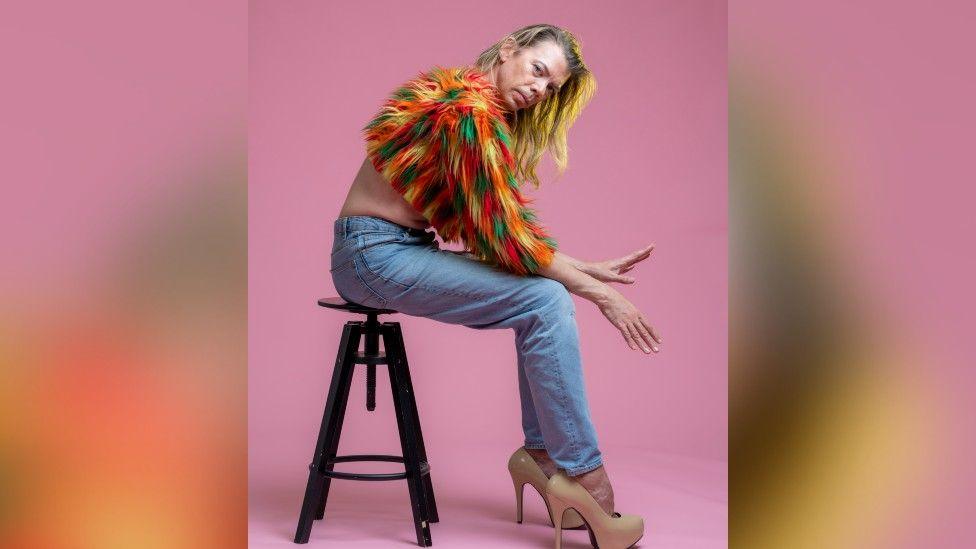 Tom Marshman wearing a multi colour feather half-jumper with denim jeans and cream stiletto heels. He is sat on a brown stool with his hands out in front, looking at the camera with a pink studio backdrop.