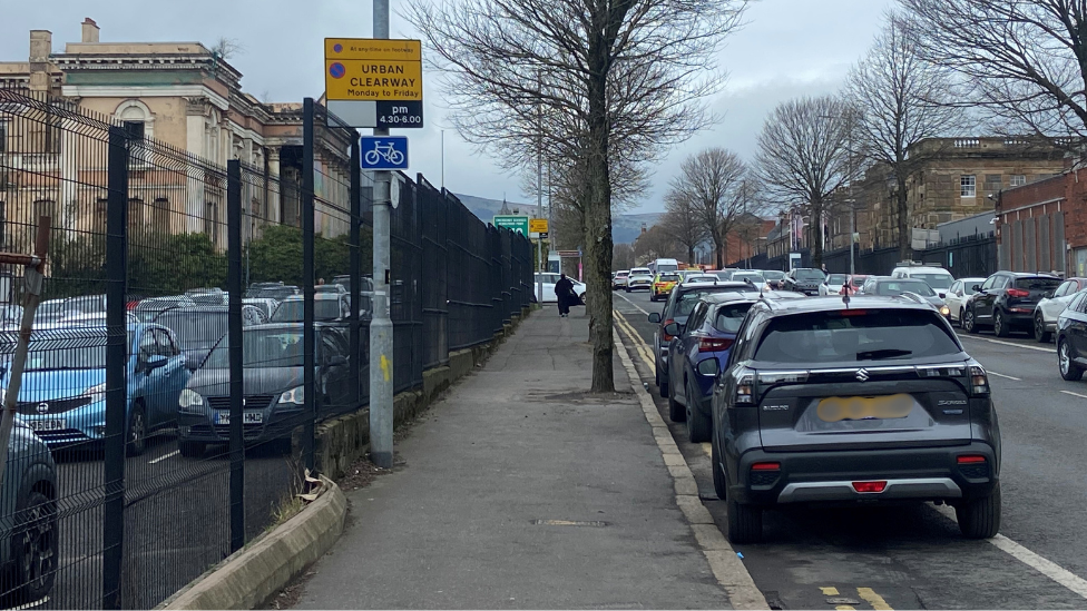 Three cars parked along a street in Belfast. A double yellow lane is on the left and a solid white line is on the right. A lamppost on the footpath has an urban clearway sign on it with a cycle sign below it.