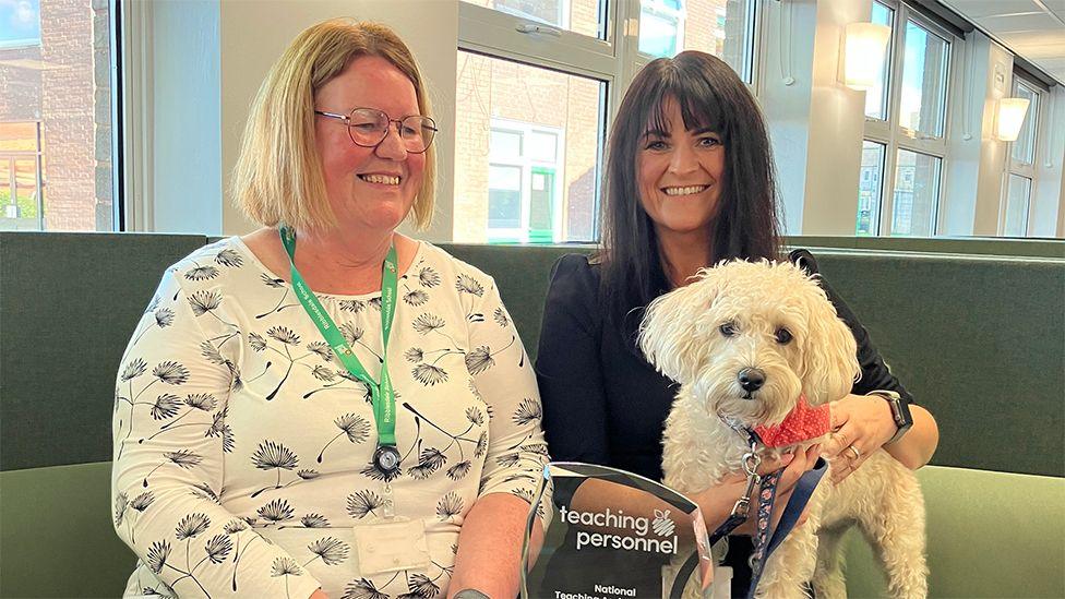 Liz Goldsbrough sitting on a green sofa next to Hannah Murphy, who is wearing black and has long dark hair and who is holding the school dog, Betty, a white curly haired cockerpoo