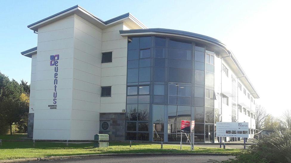 A large, white clad modern three-storey office building with glass windows curving around the main entrance and an area of grass at the front