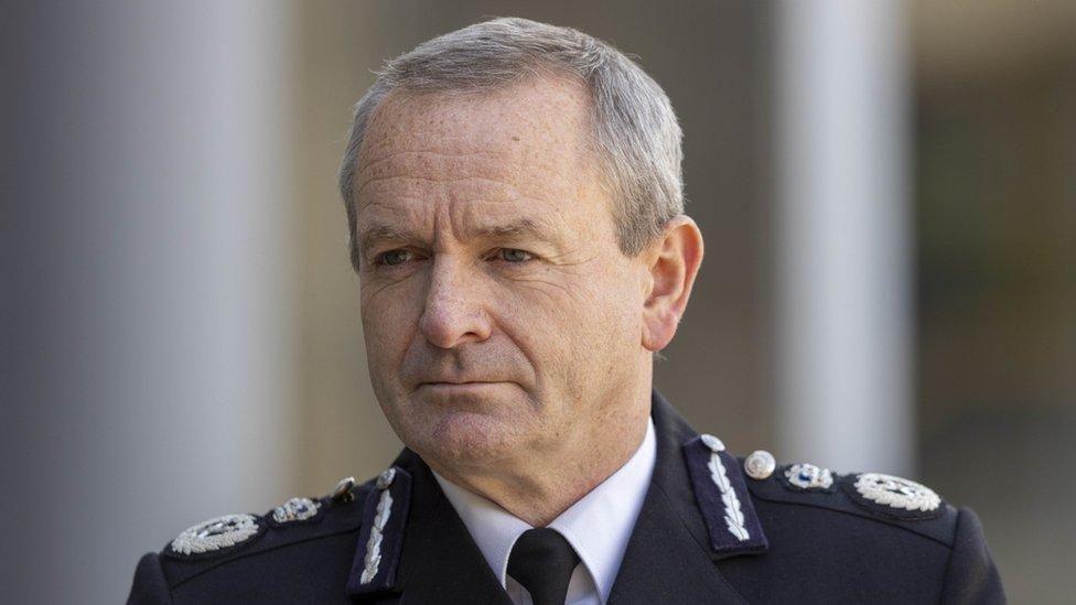 Head and shoulders shot of Sir Iain Livingstone. He is wearing his black police jacket, a white shirt and black tie. He has short grey hair, combed in a side shed and is looking off to his right with a serious expression on his face.
