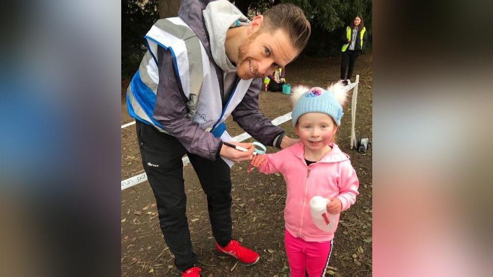 Sam is wearing a blue and silver hi-vis jacket and is behind over to hold a little girl's hand. His daughter is in a pink tracksuit set with a blue beanie on, with white fluffy bobbles. She is holding a water bottle