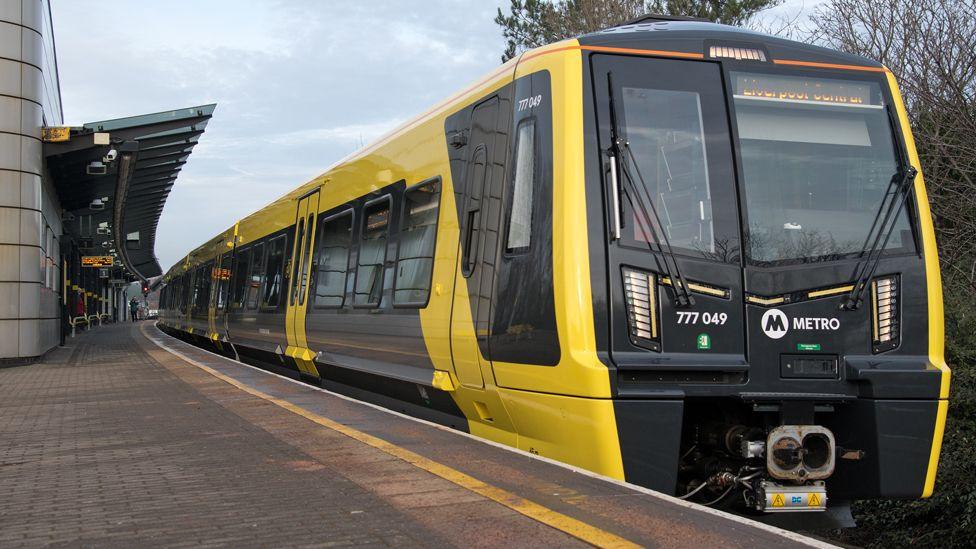 A yellow and black Merseyrail train stands at a brick-built platform, which has a grey-green awning over it and a yellow line painted close to the platform edge