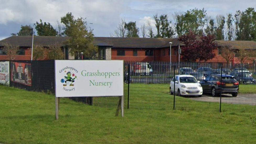 Street view image of a sig for the nursery, with a low, long, red brick building in the background and cars in a car park