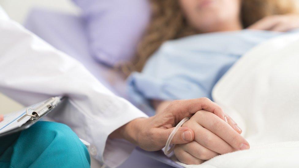 A doctor holds the hand of a woman who lies on a hospital bed