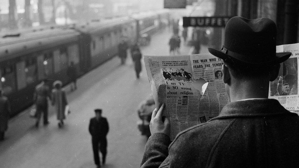 A secret agent at a train station looks through a hole in a newspaper