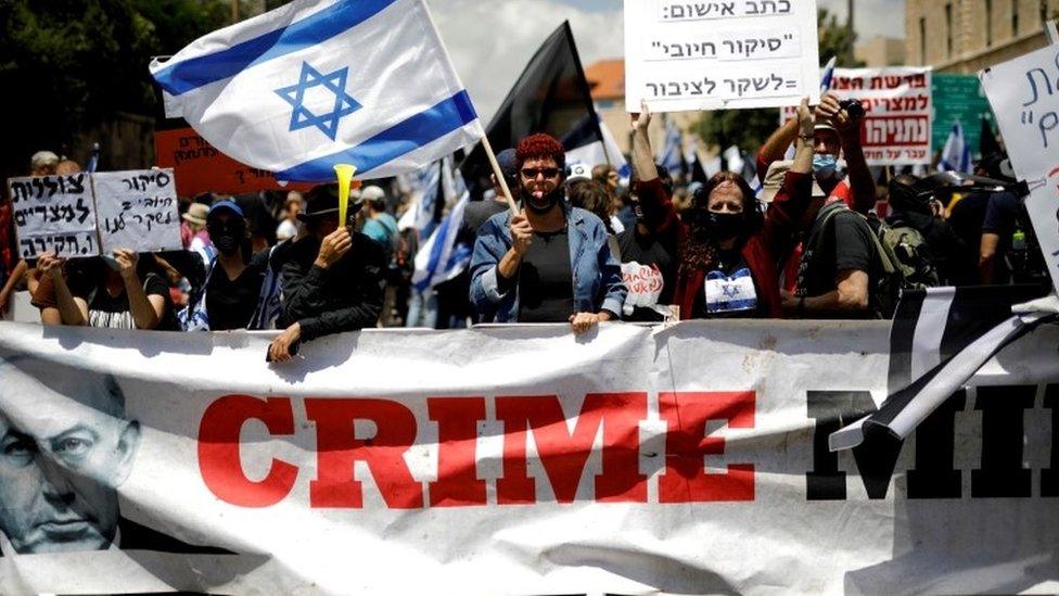 Protesters wave an Israeli flag and hold placards as they stand behind a banner reading "Crime Minister" before the start of Benjamin Netanyahu's corruption trial in Jerusalem (24 May 2020)