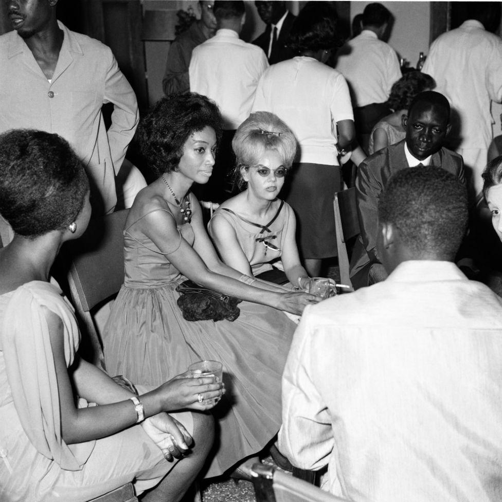 Men and women dressed in eveningwear are seen sitting down and talking with drinks and cigarettes in hand.