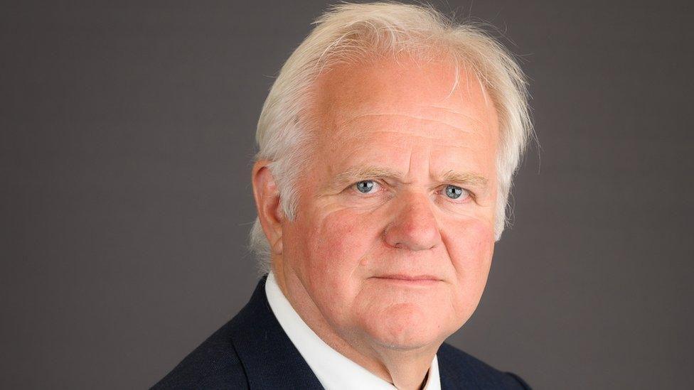 Official studio photo of councillor Mike Cox looking at the camera with a serious expression. He has white hair and is wearing a black suit with a white shirt