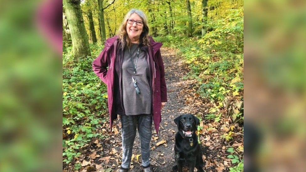 Shareen Elnagy's mum, pictured looking health and well, outside in woods with a black dog sitting by her side.