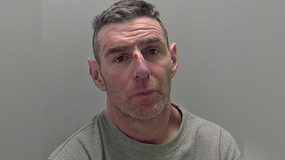 A police mugshot of a man with short dark grey hair and short grey stubble, wearing a grey sweatshirt sitting against a grey background. He appears to have an injury to his nose.