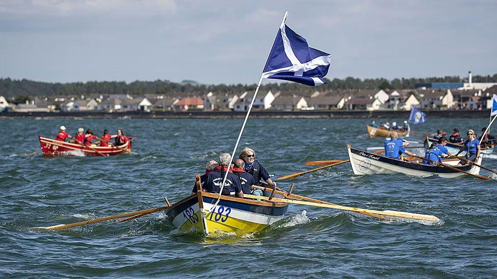 A blue, white and yellow boat with a Scotland flag on it rows out into the still waters of Loch Ryan