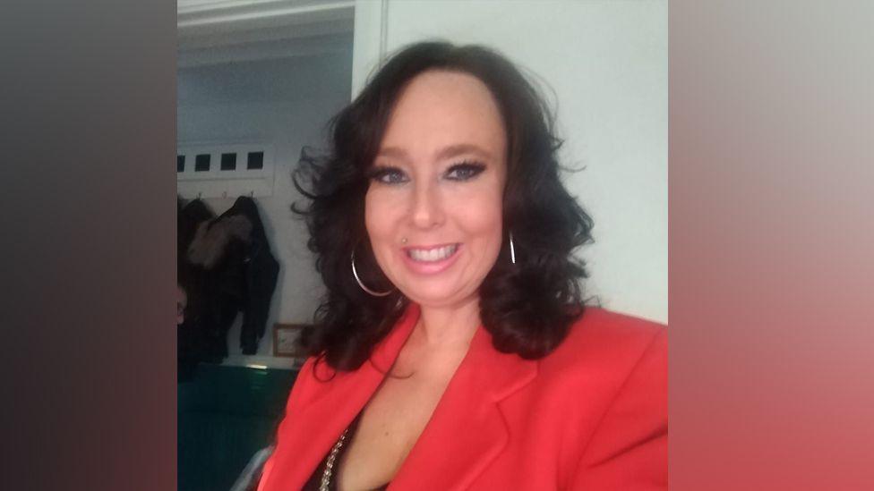 Leigh Voysey in a selfie taken wearing a red smart jacket and a black top below, She has silver hoop earrings and black wavy hair.