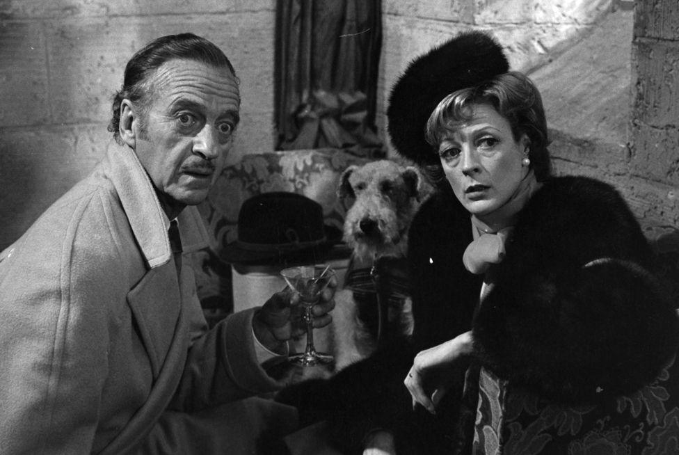 David Niven holding a martini glass as he and Maggie Smith stare forward in shock of what they see in a scene from the film 'Murder By Death', 1976