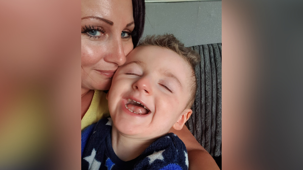 A young boy smiling, being held by his mum