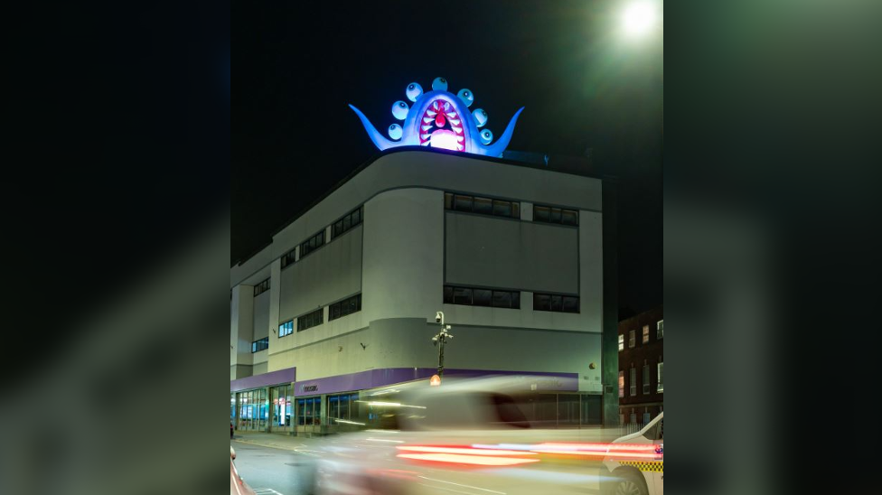 A seven-eyed blue shark-like inflatable monster on top of a block of offices. 