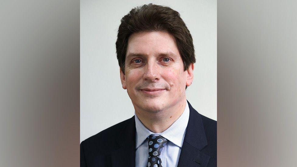 Tom Walker - a man in a black suit wearing a light blue shirt with a floral patterned black tie with blue circle flowers on it. He has dark brown hair and is smiling. White blurred background.