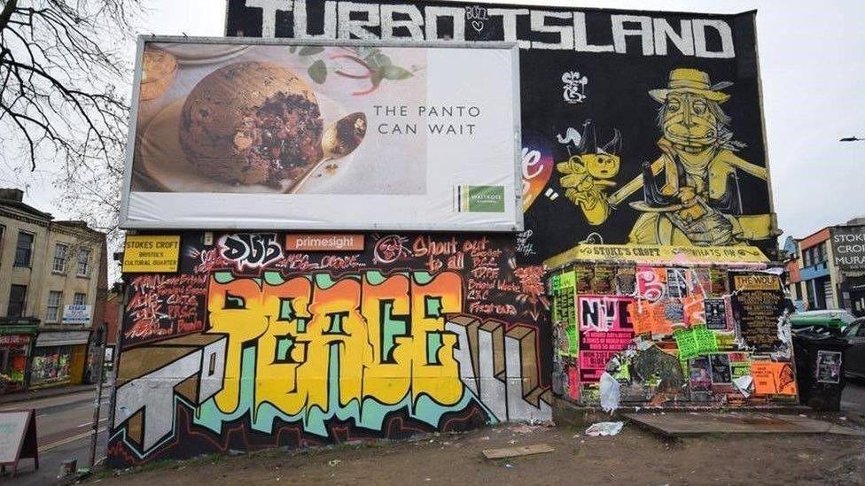 An advertising board shows a Christmas pudding, behind it is lots of graffiti. Some rubbish is left on the land in front.