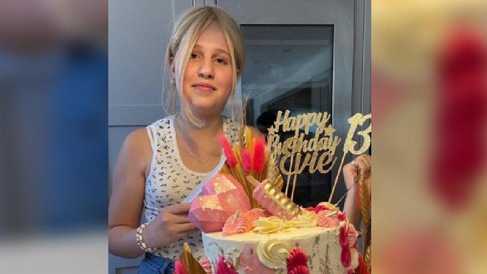 A young girl with blonde hair, wearing a white tank top with flowers on it, smiles as she stands next to a pink and white birthday cake with lots of flowers on it and a glittery gold topper that reads "Happy Birthday Evie, 13"