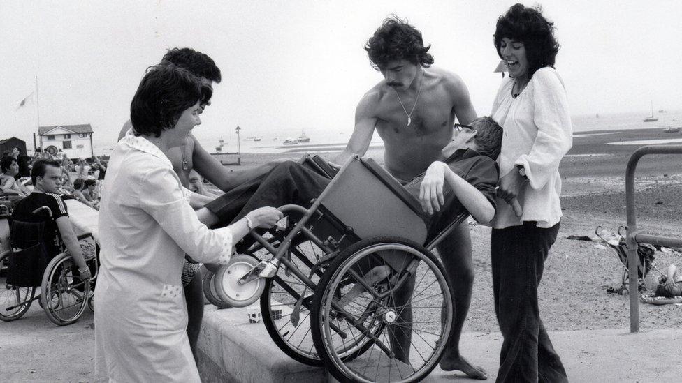 A man in a wheelchair being carried down on to the beach