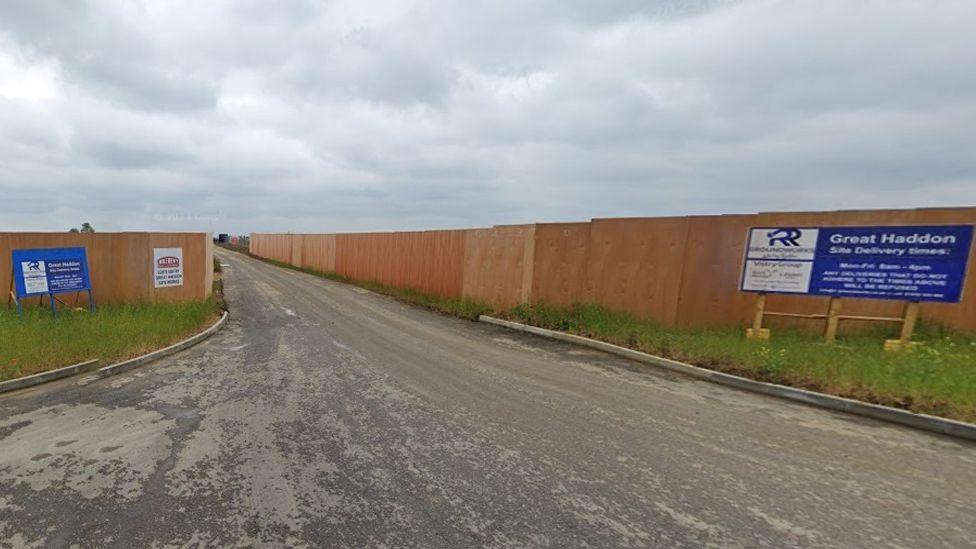 Entrance road to a building site, with tall beech coloured wooden fence panels on either side. A blue sign on the right side says Great Haddon Site Delivery times, and there is a blue notice on the left side with white writing. The grey road is rough and a bit bumpy.