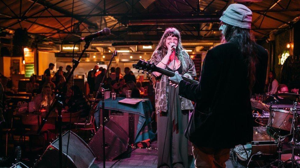 A singer sings into a microphone while a guitarist with their back to the camera plays a guitar. There are instruments on the stage and a crowd of people are standing near the bar in the background.