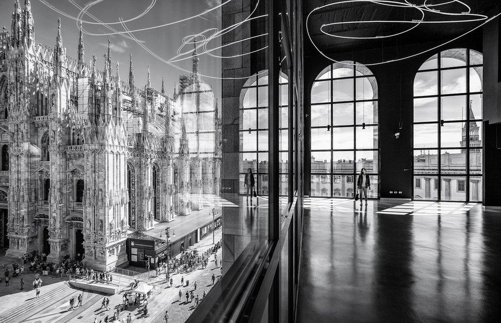 A black and white photo of the interior of a modern building next to a cathedral, with a woman reflected in glass