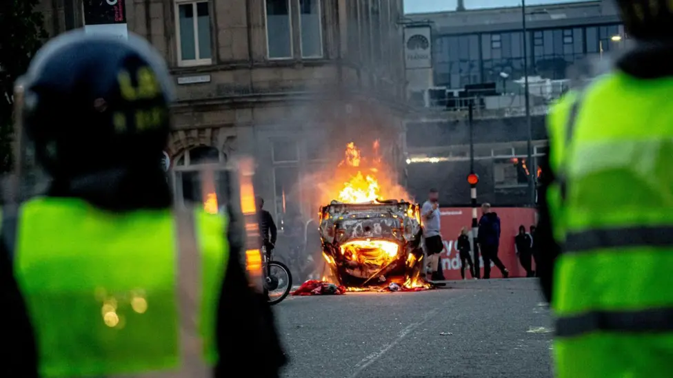 The backs of two police officers in hi vis and riot gear facing a car on fire in the middle of a street. 