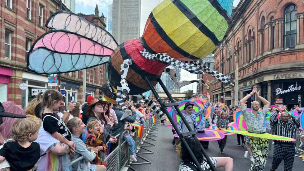 A rainbow-painted giant bee is held over a crowd of brightly-dressed spectators during Manchester Pride's parade through the city