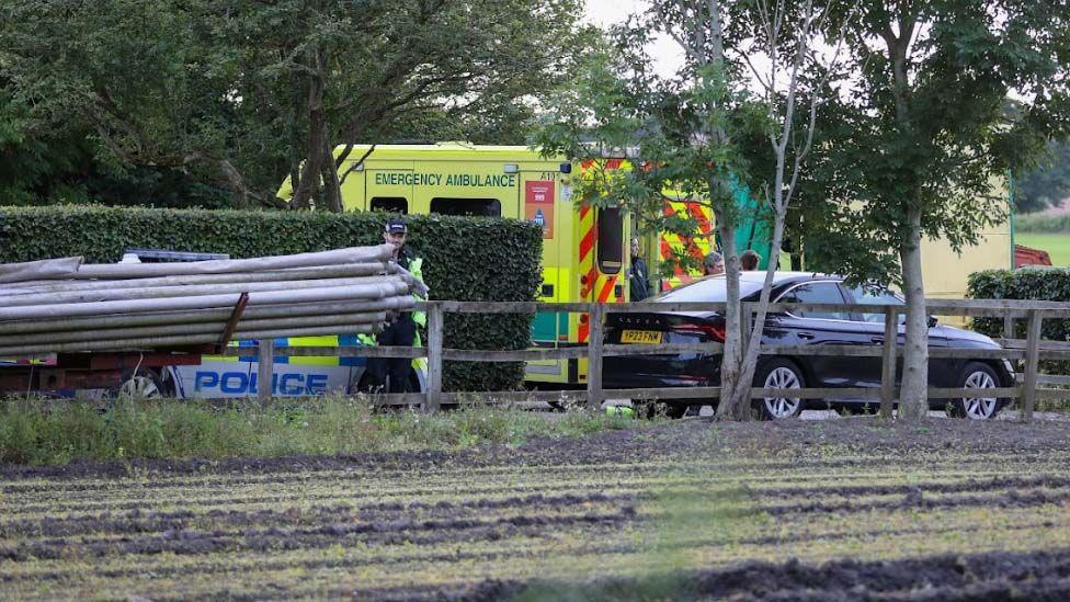 An ambulance, a police car and a black car parked next to a fence and a hedge and alongside a ploughed field, which also had trees on its borders