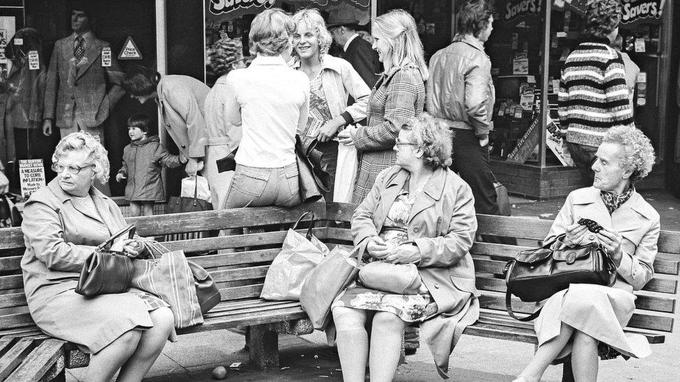 Three teenagers chatting behind a bench where three elderly ladies sit