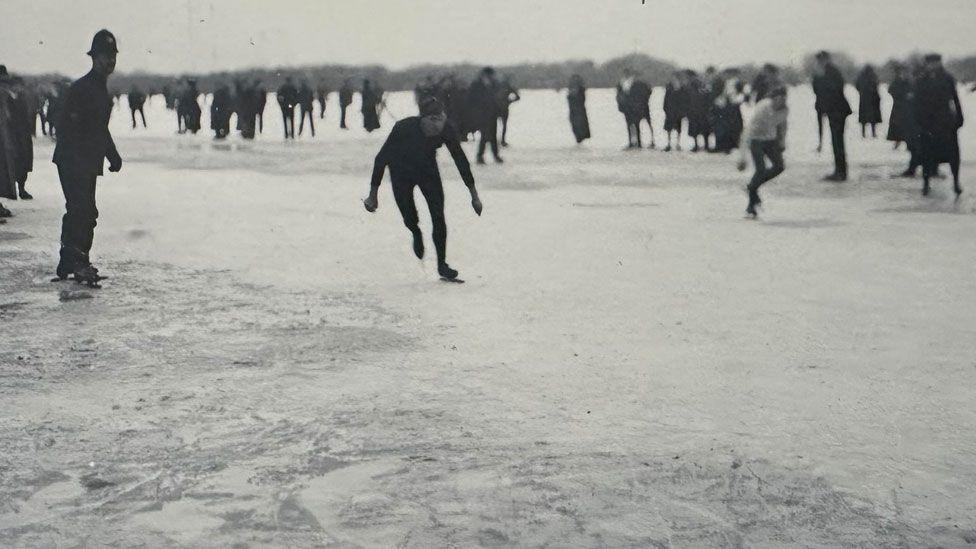 A black and white image of two skaters racing on a frozen field. Spectators can be seen behind them. On their right is a police officer, who is also on skates.