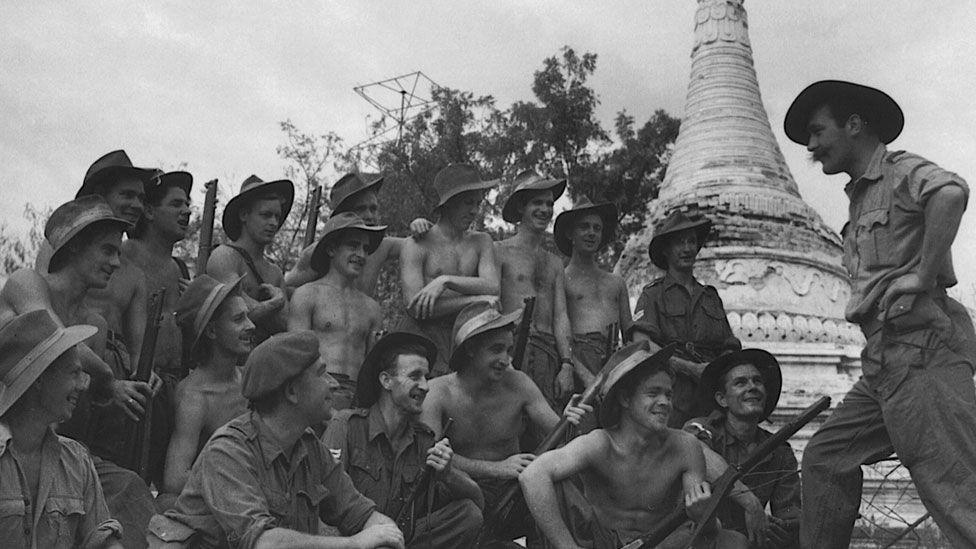 A black and white image showing a group of about 16 men from the RAF Regiment looking towards Wing Cdr Cubby Lander. The men are mostly bare-chested and many are holding guns. They are all laughing. Cubby is on the far right, side on and addressing them. He is in a field shirt and trousers, wearing a hat with his hands on his hips. Behind him in a Burmese temple tower and trees. 