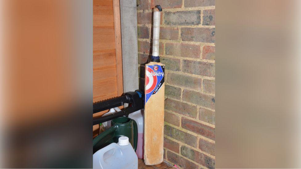 A cricket bat found near an outhouse at the family's home in Woking, It is wooden and has a white handle, with bricks in the background.