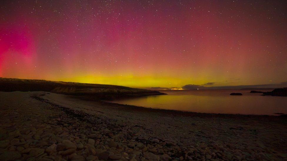 Reds and yellow colours fill the sky above the silhouette of a coastal landscape.