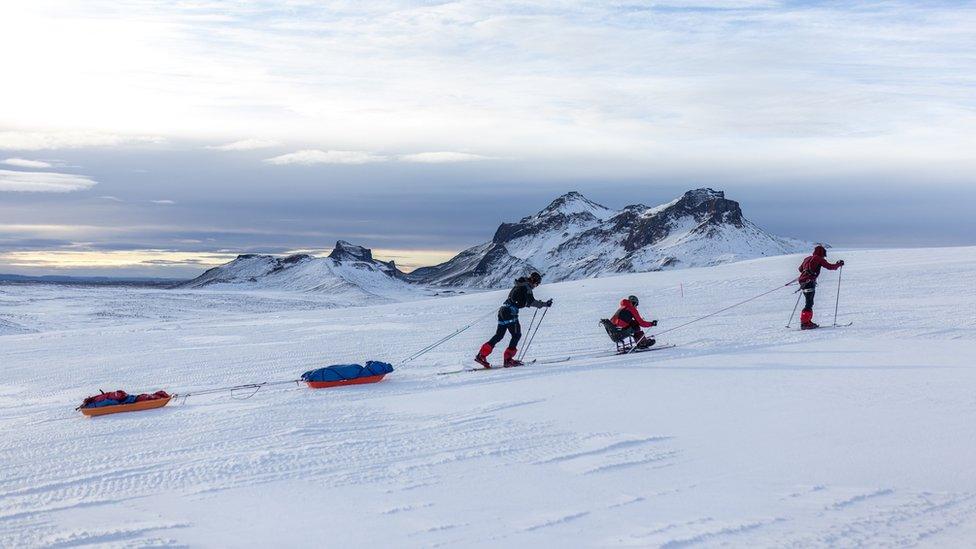 The three men skiing along a snowy mountain tied together by a rope - the middle man is on a sit ski