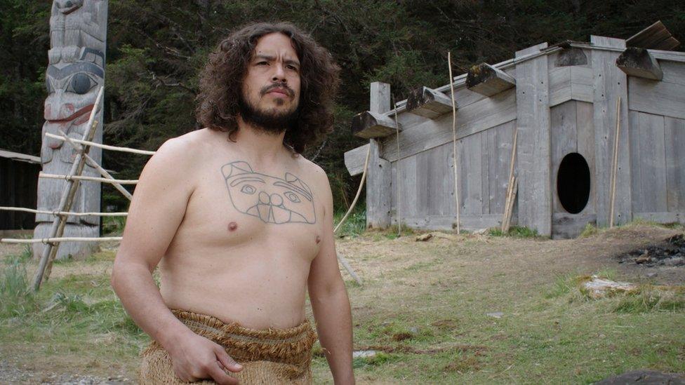 A Haida man stands in front of their traditional wooden housing