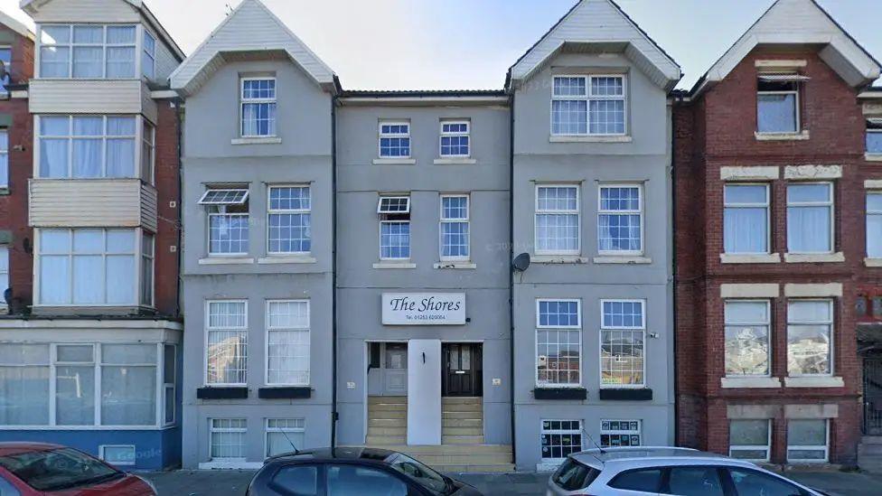 Street view image of the Shores Hotel on Tyldesley Road in Blackpool, showing  a tall grey hotel o four levels with stone steps up the front in the middle and red brick buildings to either side