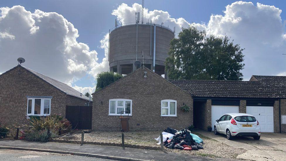 Two bungalows can be seen in front of a water tower. In front of one of the bungalows is a pile of discarded items