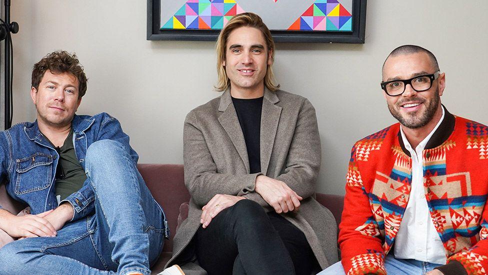 James Bourne, Charlie Simpson and Matt Willis from the band Busted sitting together on a sofa smiling in casual clothing