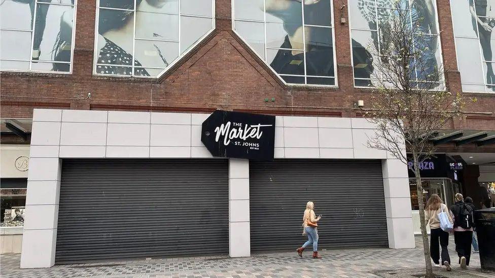 A woman walks past the external entrance to St Johns Market in Liverpool which has two black shutters down. A sign reads The Market, St Johns in white writing on a black background.