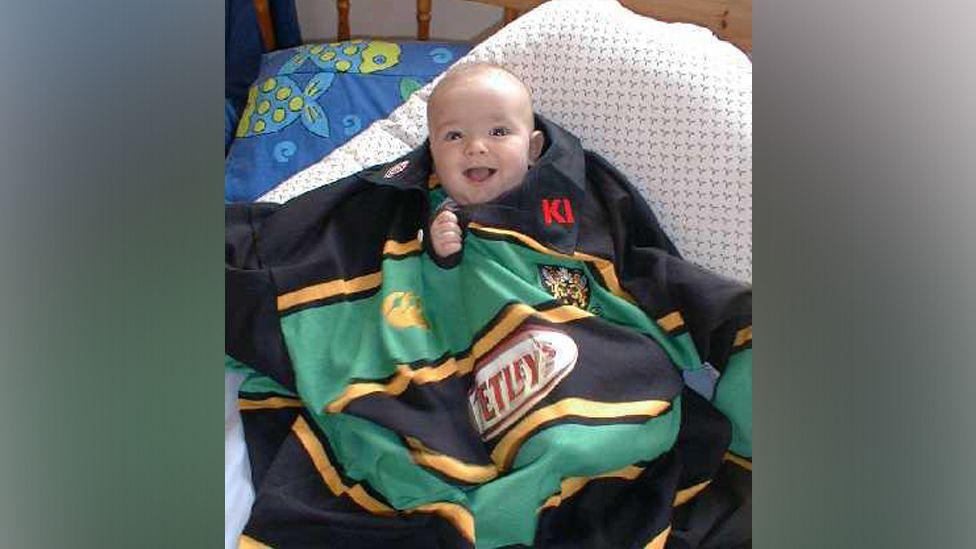 Matthew Sansome as an infant. He is on a bed and has a black, green and gold Northampton Saints jersey draped over him, with his head and one hand peeping out of the collar.