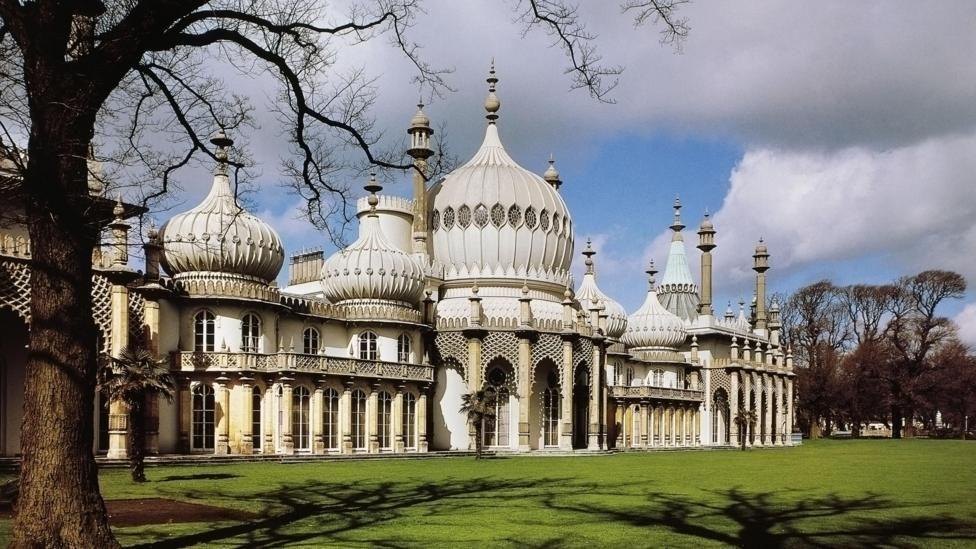 Brighton's Royal Pavilion, a large white palatial building with domes rooftops resembling onions. It is surrounded by a neat grass lawn and trees.