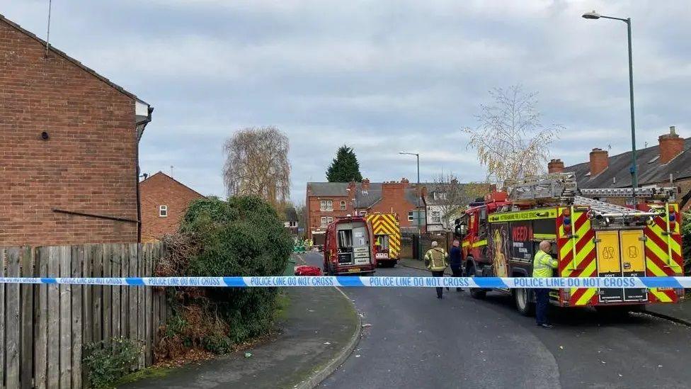 Police tape strung across a street with fire crews parked up behind it
