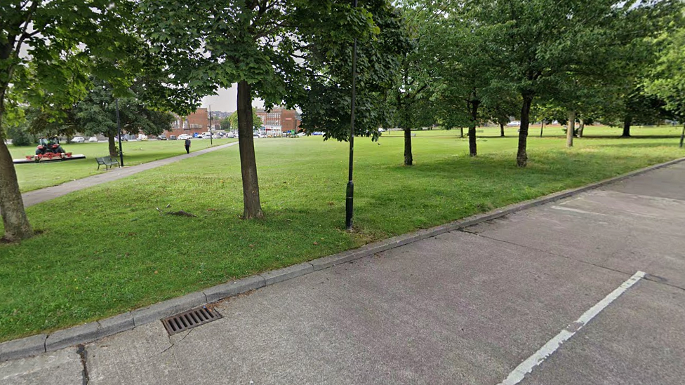 A wide shot of a grassy square ringed with trees with a path going through the centre of it